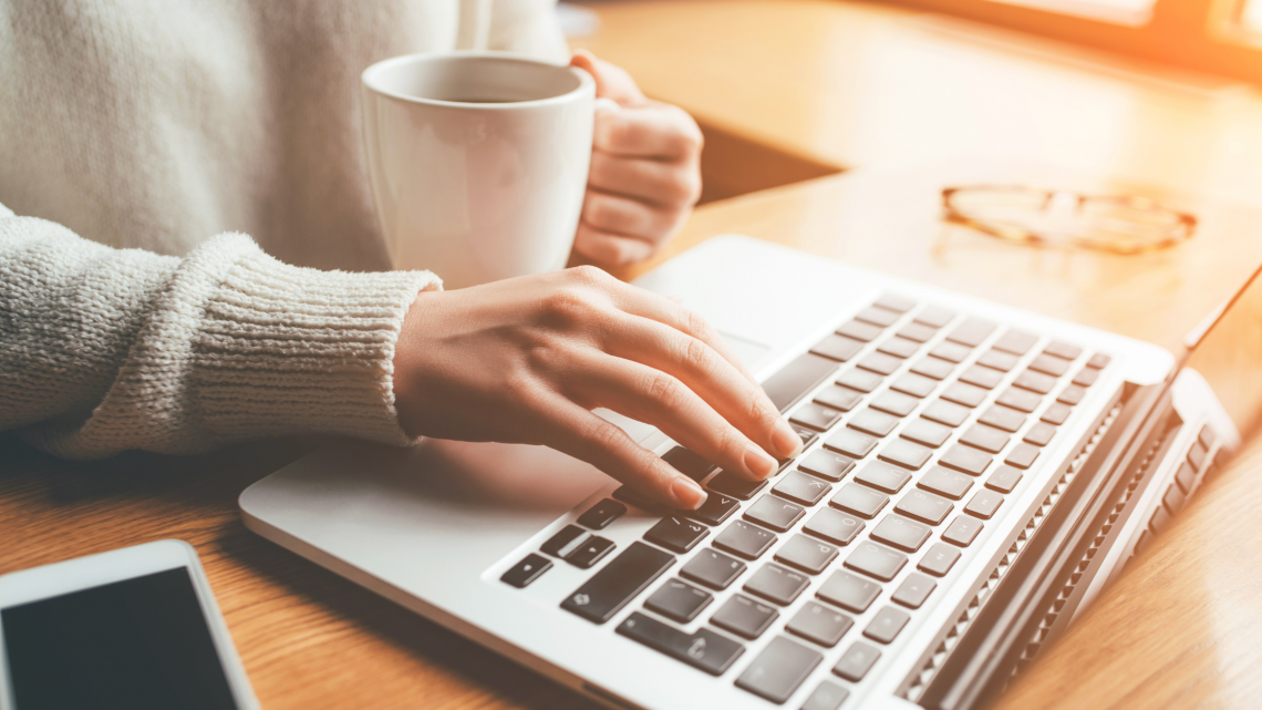 Photo of a woman's hands holding a coffee mug and typing on a laptop