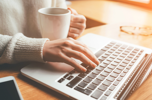 Photo of a woman's hands holding a coffee mug and typing on a laptop