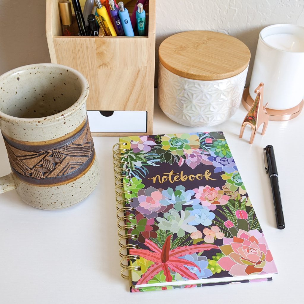 Photo of a journal, pen, coffee mug, and candle on a white desktop