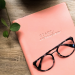 Photo of a pink journal, plant, and glasses sitting on a wooden tabletop